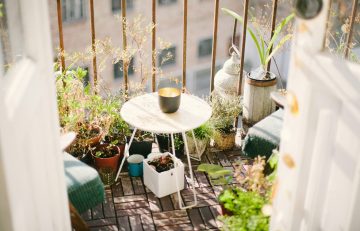 Kleiner, sonniger Stadtbalkon mit Holzboden, einem runden Beistelltisch, Topfpflanzen und gemütlicher Sitzgelegenheit – liebevoll gestaltet mit grünen Pflanzen, Kräutern und einer Laterne.